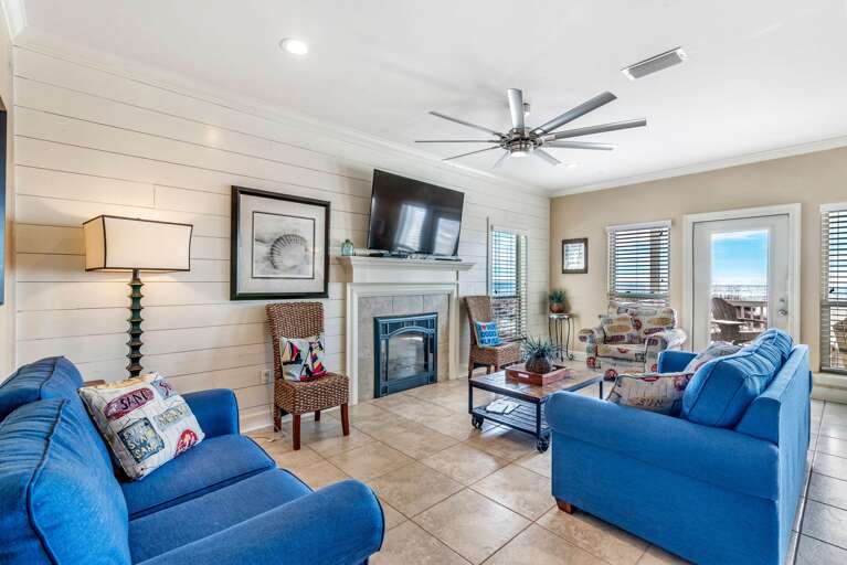 Tiled living room floor with blue couches, patterned armchairs, shiplap wall, and glass door framing ocean view.