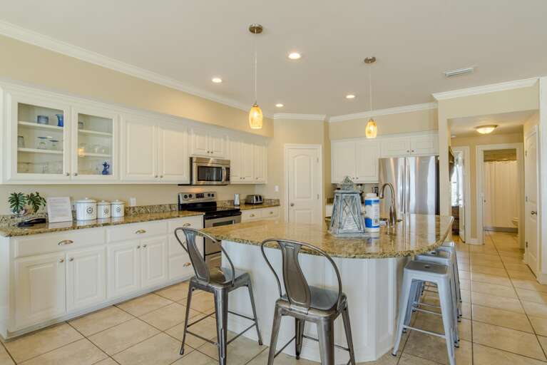 Kitchen features granite countertops, white cabinetry, stainless steel appliances, and metal barstools under amber pendant lights.