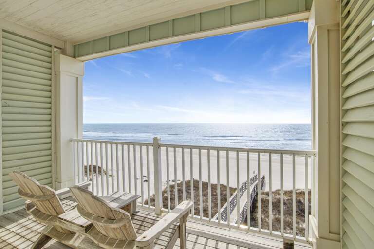Covered balcony with weathered wood Adirondack chairs and white railing overlooking sandy beach and ocean.