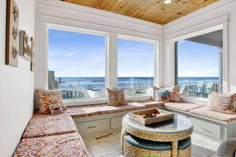 Sunlight fills the corner living room nook with patterned cushions and a wicker table, overlooking ocean waves.