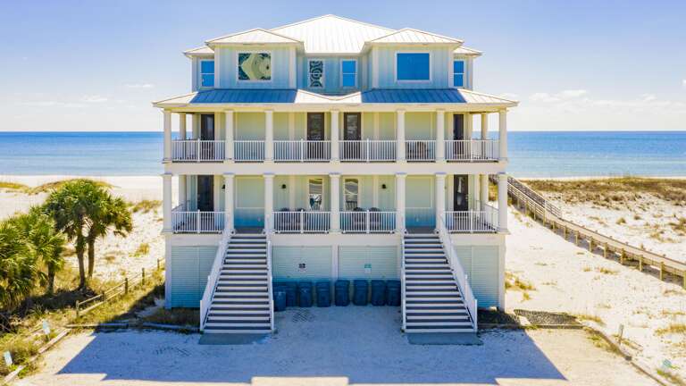 Beachfront exterior of a three-story white house with twin staircases and wraparound balconies overlooking the Gulf.