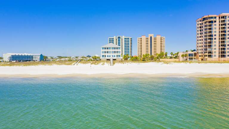 Clear waters ripple toward a wide, sandy beach with a three-story blue beach house centered among taller buildings.