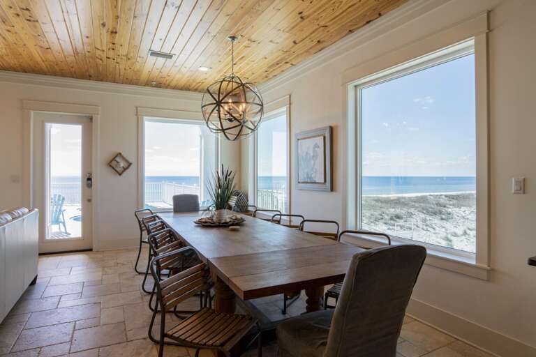 Dining area with large wooden table surrounded by metal and upholstered chairs, natural wood plank ceiling, and ocean views.