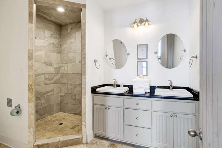 Bathroom with dual rectangular sinks set in black granite countertop, round mirrors, and a tiled walk-in shower.