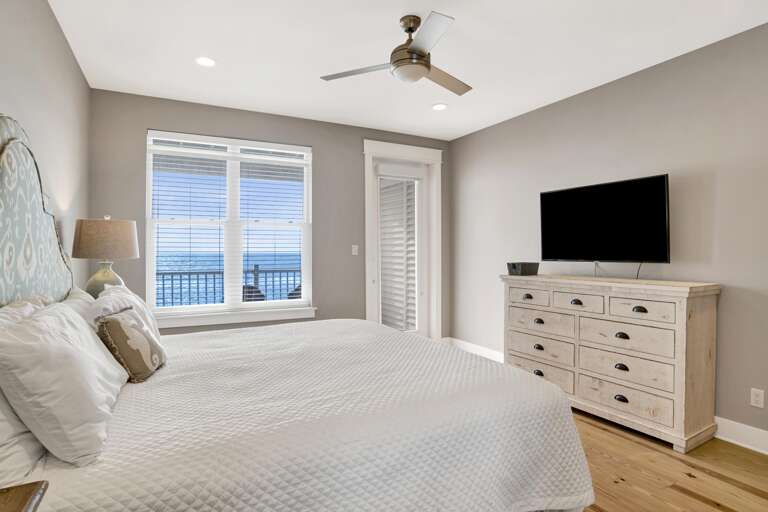 King bed with patterned blue headboard faces a whitewashed wood dresser and wall-mounted TV, ocean visible through blinds.