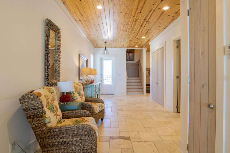 Living room entry with beige stone tile flooring, wicker armchairs with sea-themed cushions, and pine wood ceiling.