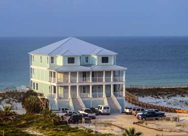 Three-story beach house with pastel green siding, metal roof, and wraparound balconies facing the gulf.