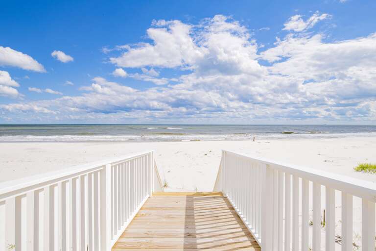 Boardwalk with white railings leads down to the white sandy beach and gentle waves under a partly cloudy sky.