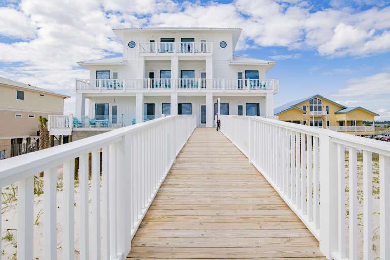 Wide wooden boardwalk with white railings leads to a three-story white beach house with multiple balconies and circular windows.