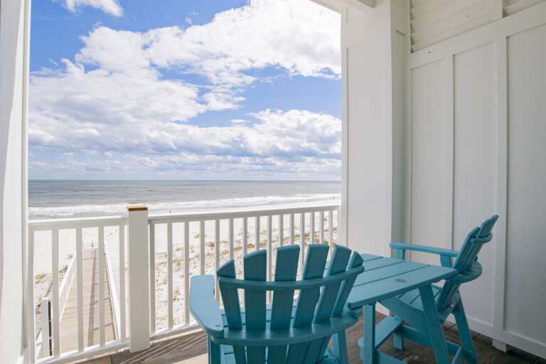 Turquoise Adirondack chairs face the ocean from a white-paneled balcony with wooden plank flooring.