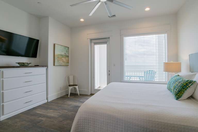 Bedroom with white quilted bedding, wood-look tile floor, wall-mounted TV, and glass door to balcony.