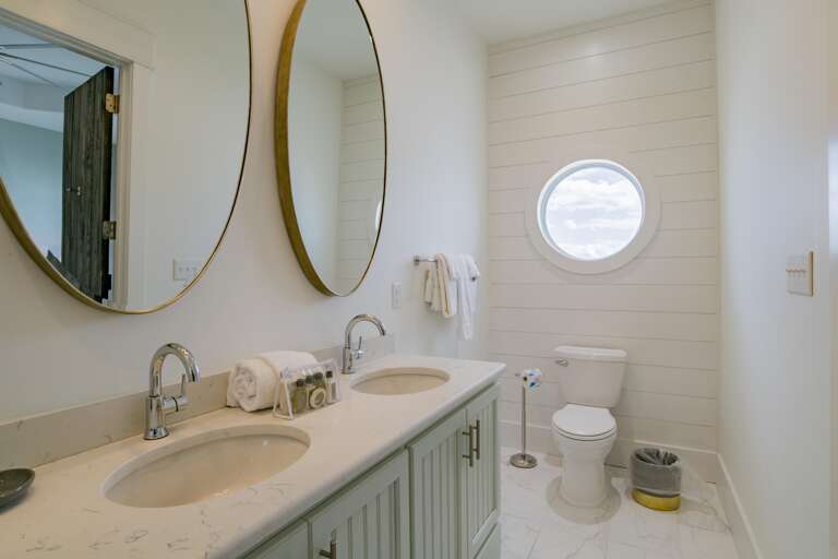 Double vanity with marble countertop and oval mirrors in a white shiplap bathroom featuring a round porthole window.