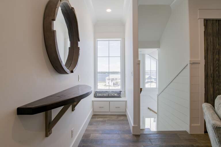 Narrow hallway with dark wood shelf and round mirror, leading to a window seat overlooking the water.