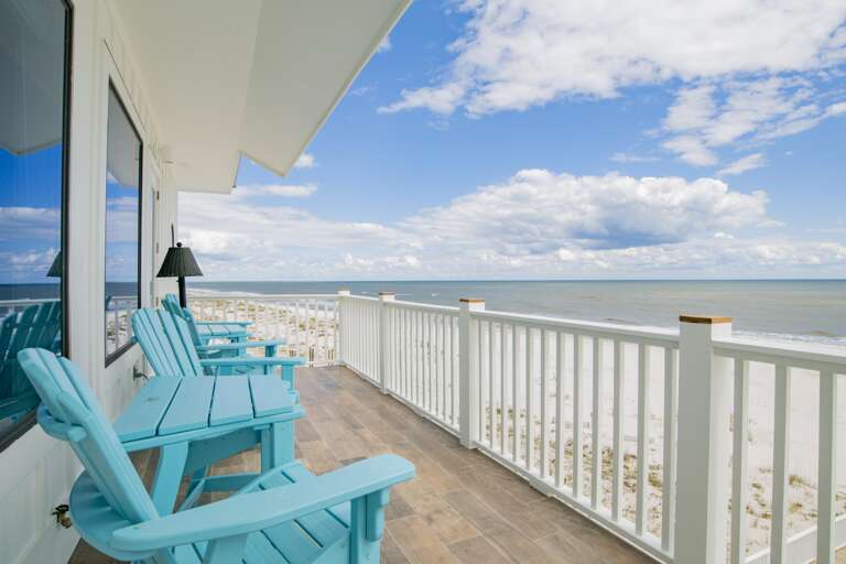 Balcony with wood-look tile flooring, white railing topped with natural wood, and turquoise Adirondack chairs facing the beach.