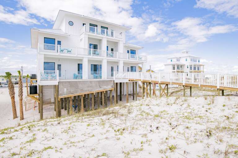 Three-story white beach house on wooden stilts with multiple balconies and a wide boardwalk over sandy dunes.