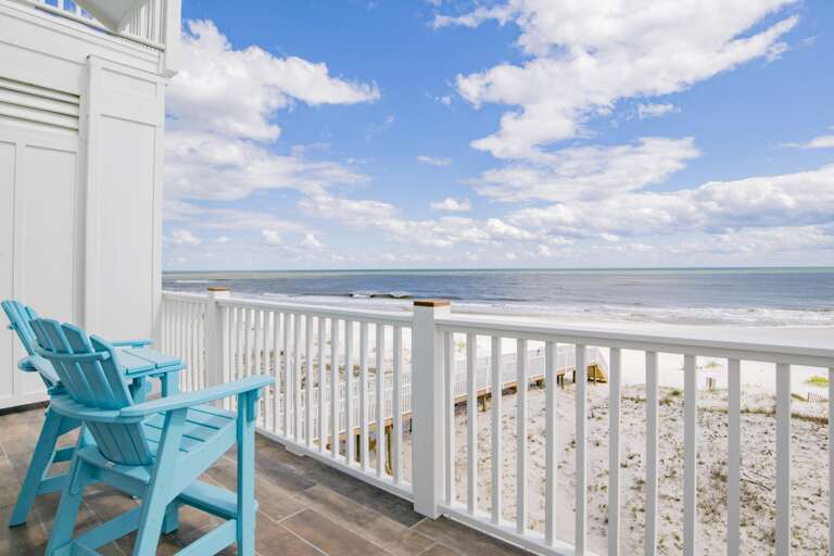 Balcony with blue Adirondack chairs overlooks sandy dunes and the calm Gulf waters under a partly cloudy sky.