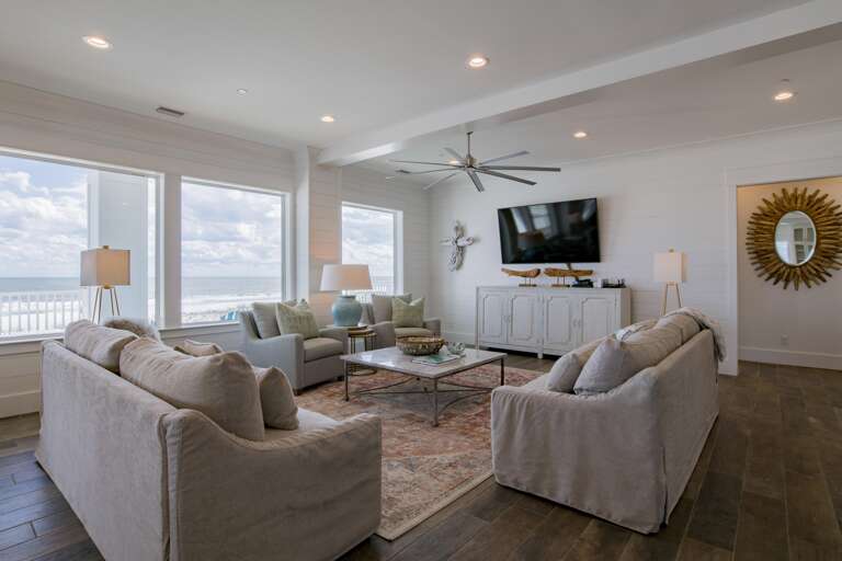 Expansive windows frame ocean views behind beige sofas and armchairs centered around a patterned area rug and metal coffee table.
