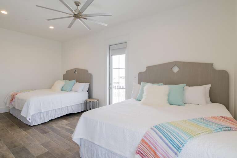 Two queen beds with gray wood headboards, white linens, pastel pillows, and a door leading to a balcony.