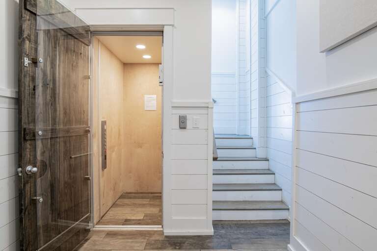 Open elevator with light wood interior next to white shiplap staircase and rustic wood-framed glass door.