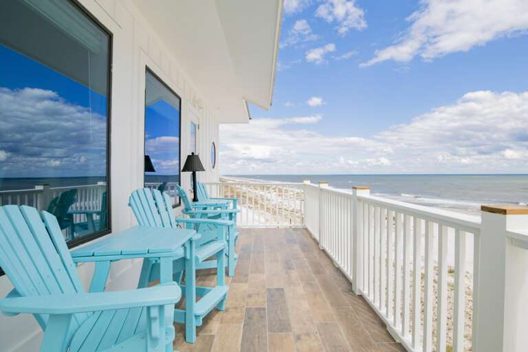 Oceanfront balcony with wood-look tile floor, white railings, and aqua Adirondack chairs facing the beach.