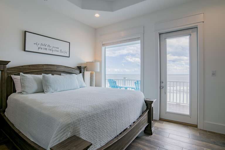 King bed with weathered wood frame faces sliding glass door and window opening to balcony and ocean views.