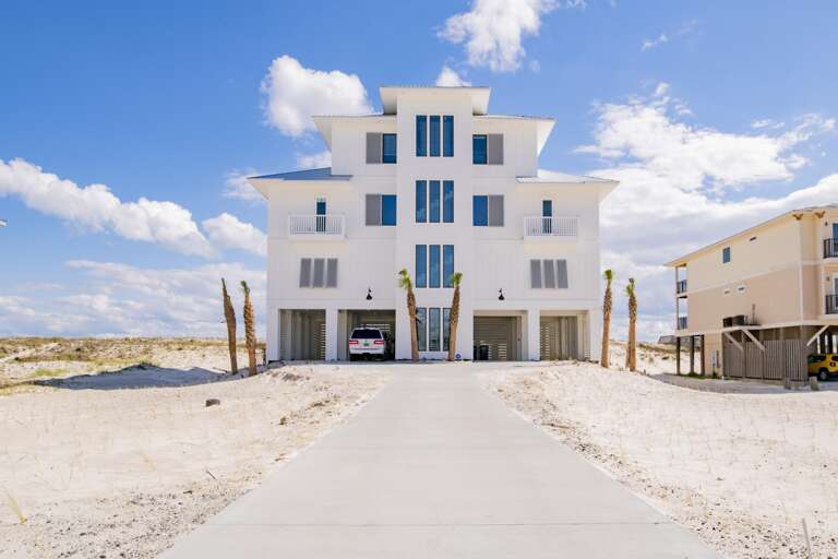 Front view of the three-story white Gulf-front house with metal roof, tall windows, and a concrete driveway.