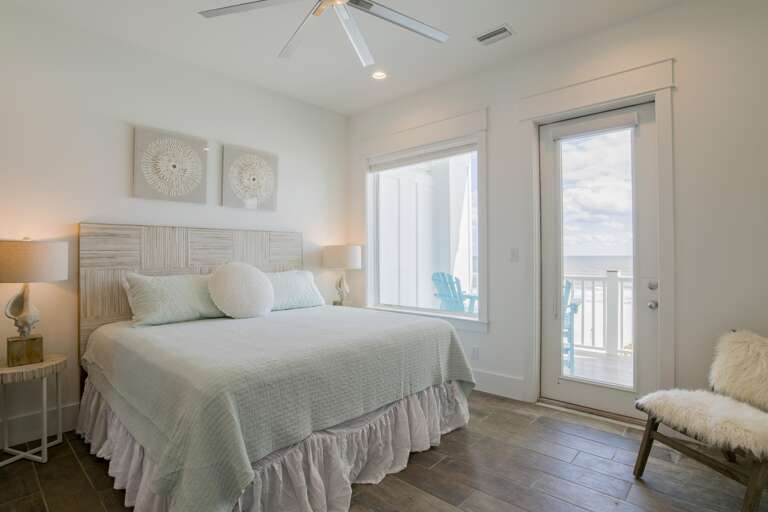 Bedroom with pale wood headboard, sea-inspired wall art, tiled flooring, and glass door opening to ocean balcony.