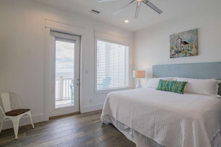 Bedroom with white linens, light blue headboard, wooden floors, and glass door opening to balcony ocean view.