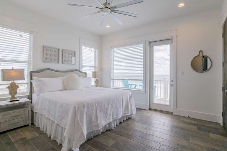 Bedroom with wood-look tile flooring, white bedding, twin lamps on driftwood nightstands, and balcony access with ocean views.