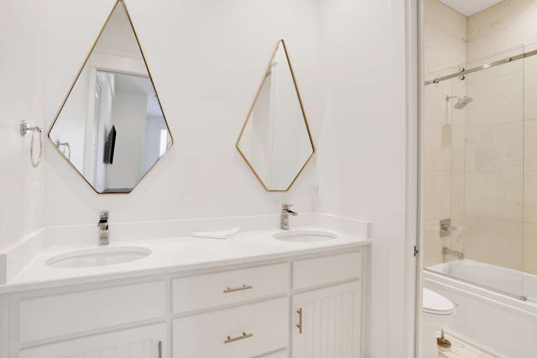 White dual-sink vanity with geometric mirrors and a glass-enclosed tub with beige tile surround.