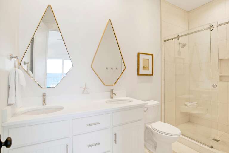 Bathroom features dual white vanities beneath angled gold-framed mirrors, a glass-enclosed tiled shower, and a white toilet.