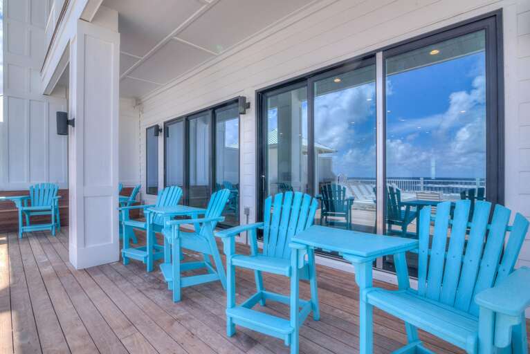 Covered balcony with turquoise Adirondack chairs and small tables on natural wood planks, reflecting ocean views.