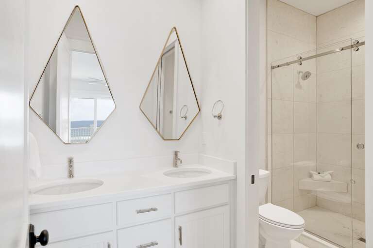 Bathroom with dual white vanities beneath gold-framed diamond-shaped mirrors and a tiled glass shower enclosure.