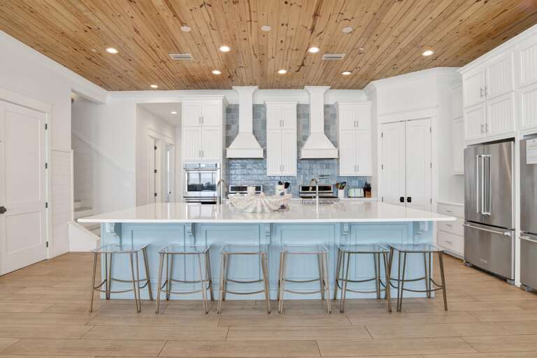 Kitchen island with pale blue cabinetry and six metal-framed stools beneath a knotty pine ceiling and recessed lighting.