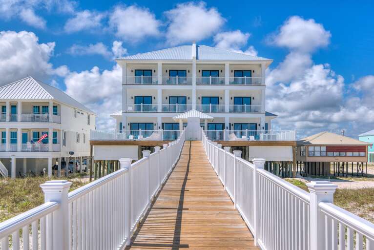 Wood-plank boardwalk with white railings leads to the raised, white three-story beachfront house under a bright blue sky.