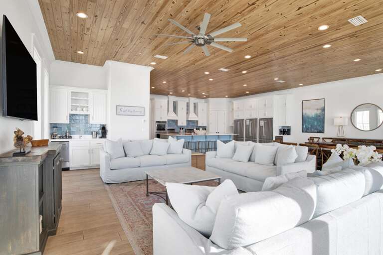 Three white sofas surround a square coffee table on a patterned rug beneath a wide pine ceiling fan.