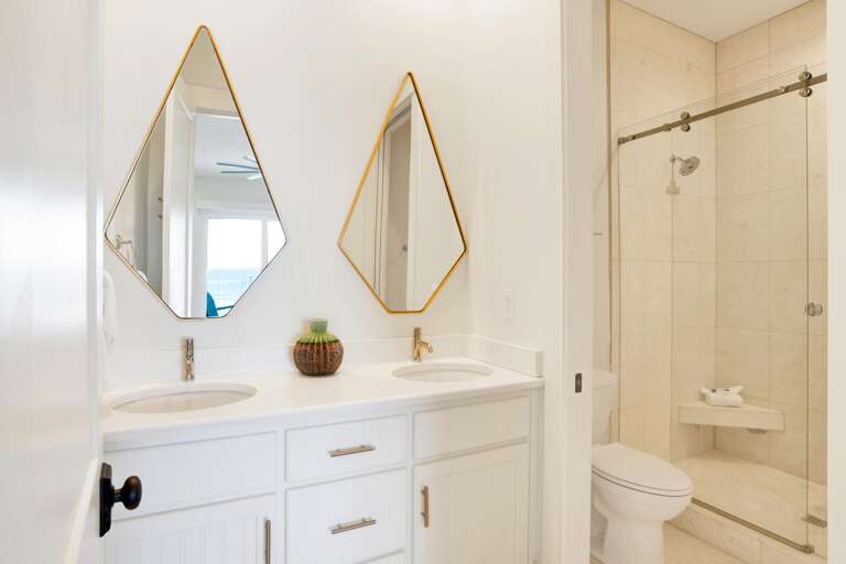 Bathroom with dual white vanities, teardrop mirrors with brass frames, and a glass-enclosed beige tile shower.