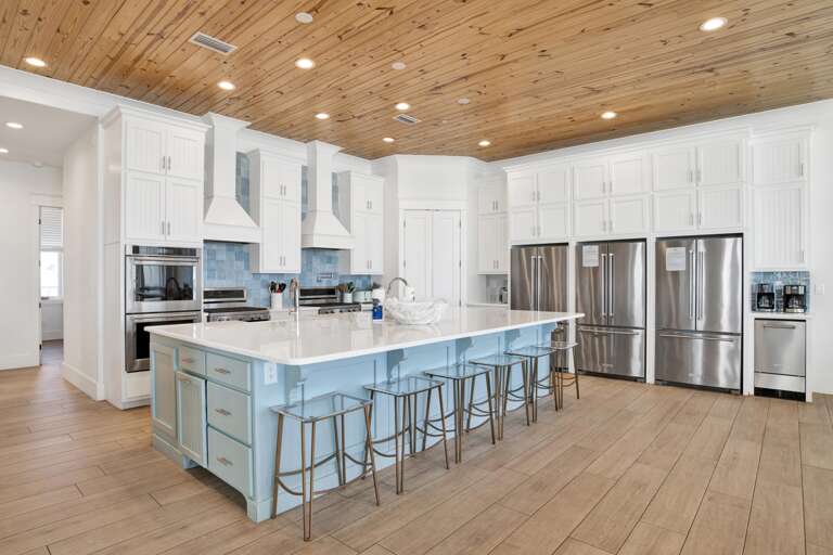Kitchen island with pale blue cabinetry, six metal stools, and white quartz countertop beneath a knotty pine ceiling.