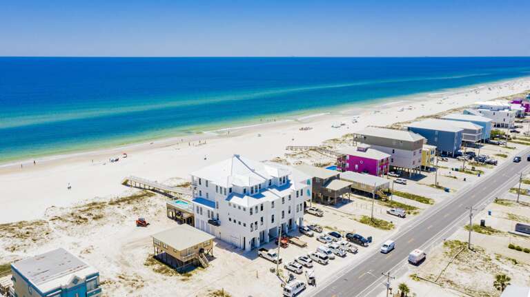 Oceanfront view from above showing white three-story house, sandy beach, and turquoise Gulf waters stretching to horizon.