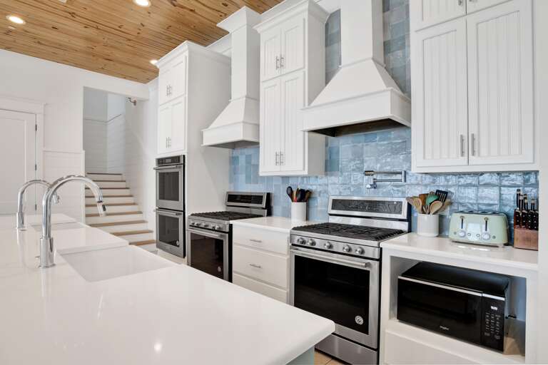 Kitchen with dual stainless steel ovens and gas ranges, glossy blue tile backsplash, and white cabinetry under a pine ceiling.