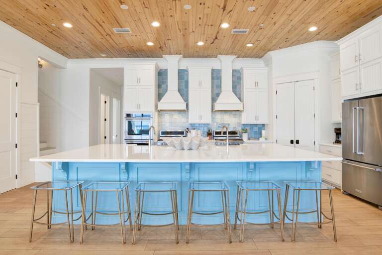 Blue island with white quartz countertop and six clear acrylic stools in the kitchen under a knotty pine ceiling.