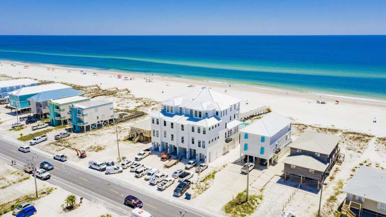 Wide aerial shot of the beachfront house with white metal roof, multiple windows, and sandy shoreline stretching into the horizon.