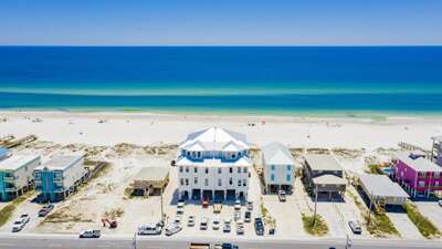 Aerial view of the white three-story beachfront house with metal roof, multiple windows, and parking spaces facing the Gulf.