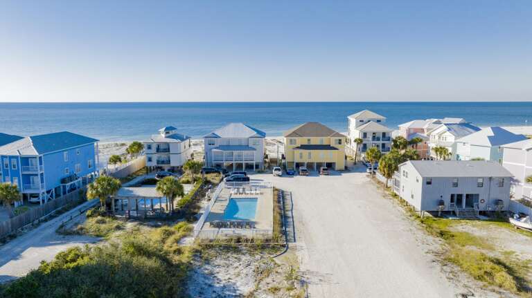 Aerial view of beachfront homes with a fenced pool, palm trees, and clear blue Gulf waters in the background.