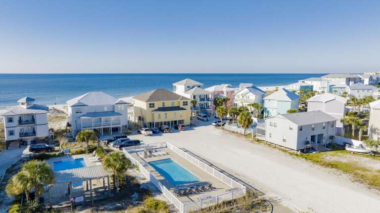 Two rectangular fenced pools with lounge chairs sit near palm trees and sandy beachfront homes under a clear blue sky.
