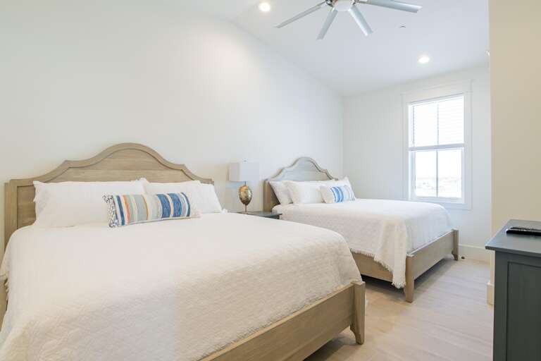 Two queen beds with light wood frames and white bedding sit under a ceiling fan by a window with blinds.