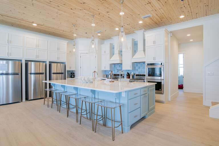 Kitchen with pale blue island, quartz countertop, six metal stools, stainless steel appliances, and pine plank ceiling.