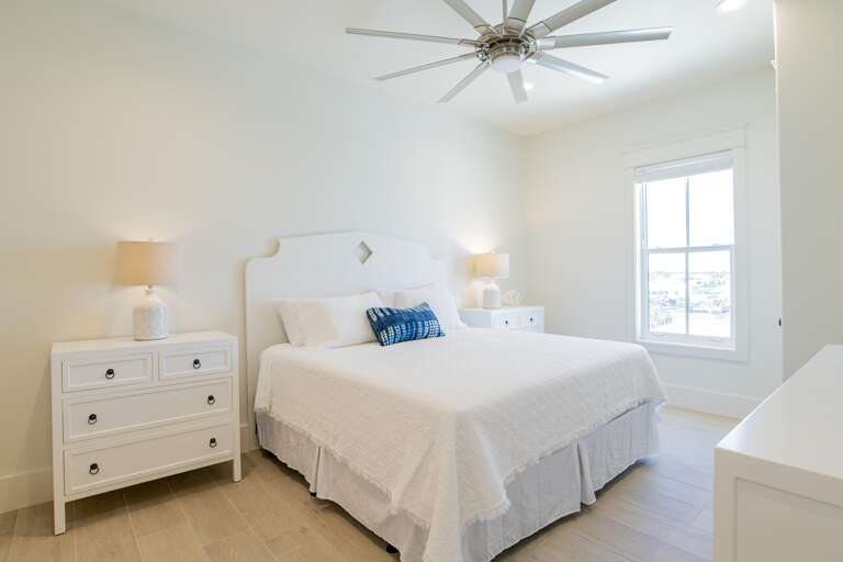 Bedroom with white textured bedding, matching nightstands with lamps, light wood floors, and window view.