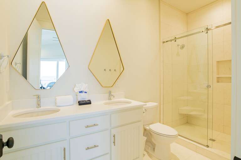 Bathroom with dual sinks, diamond-shaped mirrors, white cabinetry, and glass-enclosed tiled shower with built-in bench.