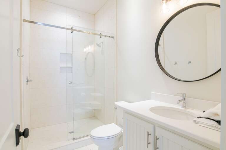 Bathroom with white quartz countertop, round black-framed mirror, and glass-enclosed walk-in shower with built-in bench.